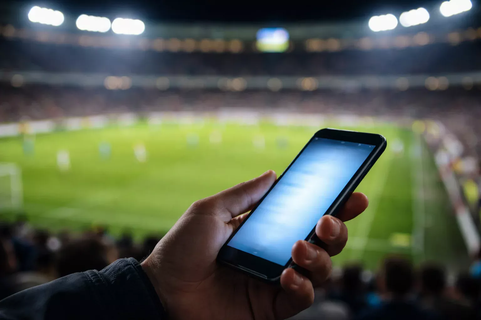 Mano sosteniendo un teléfono móvil con la pantalla iluminada frente a un campo de fútbol nocturno